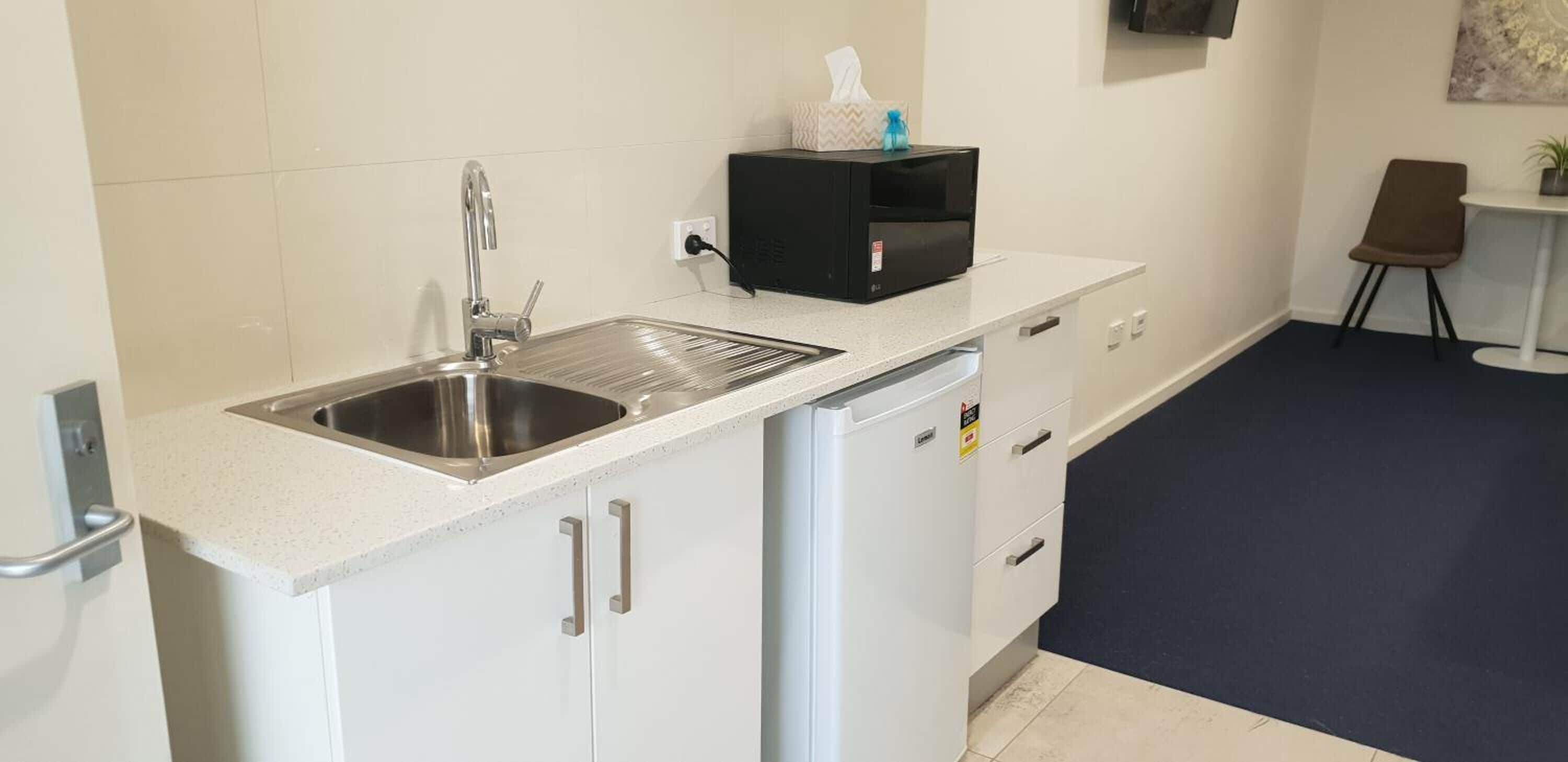 A kitchenette with a stainless steel sink, a microwave, and a small refrigerator. There is a chair and a table in the adjacent area.