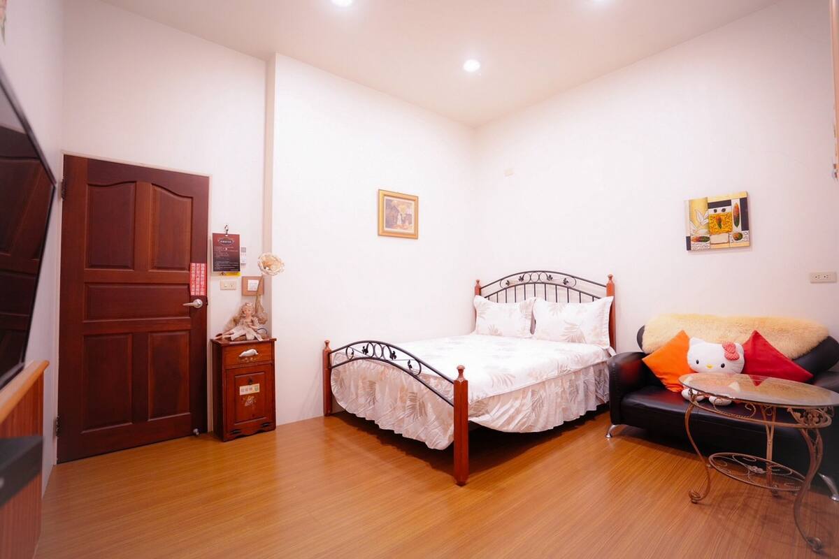 A bedroom with a wooden floor, a bed with a metal frame, a small bedside table, a sofa with a cushion, and a wall-mounted picture.