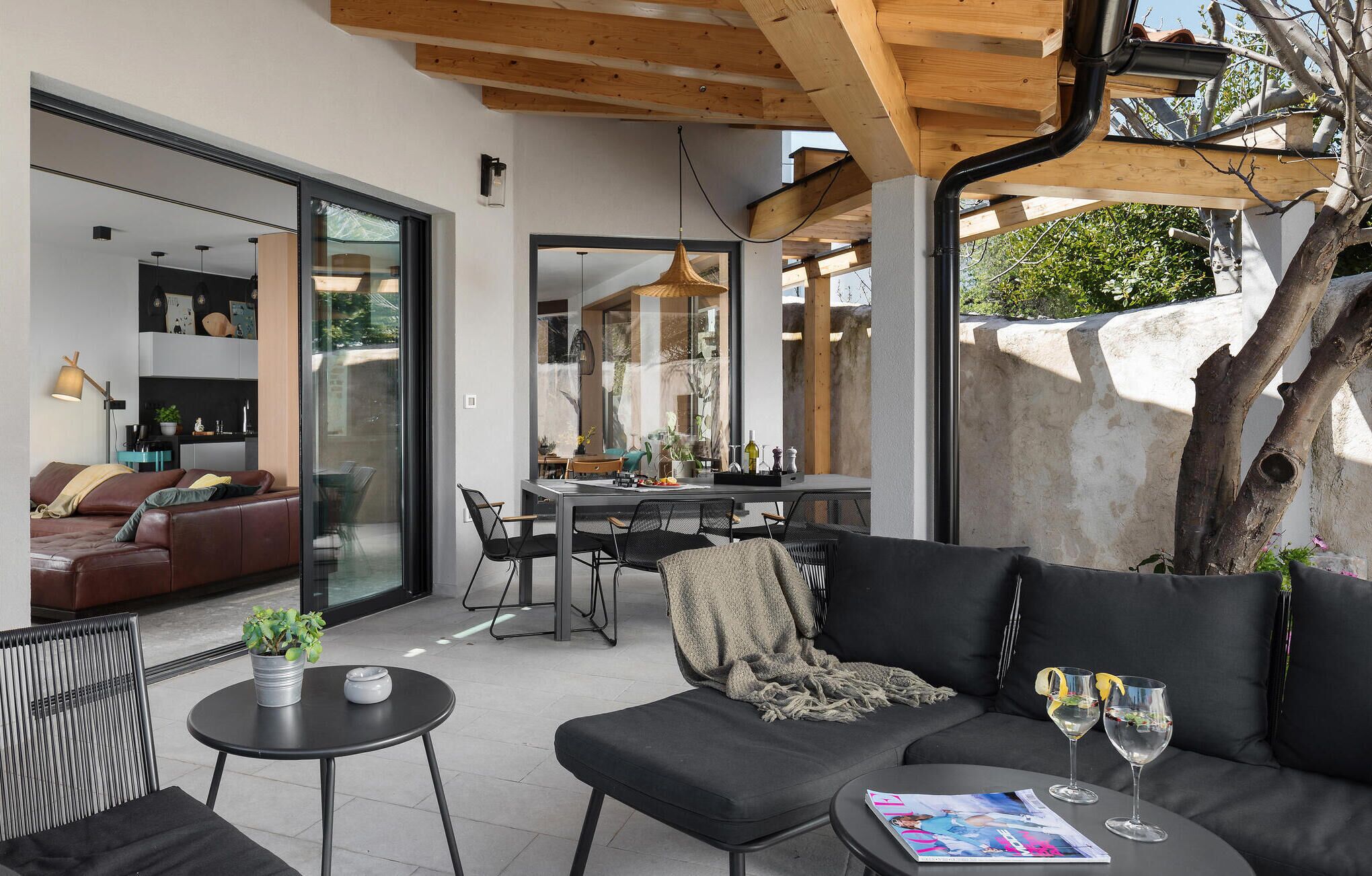 A modern living room with a sofa, a dining table, and chairs. There is a large window providing a view of the outdoors.