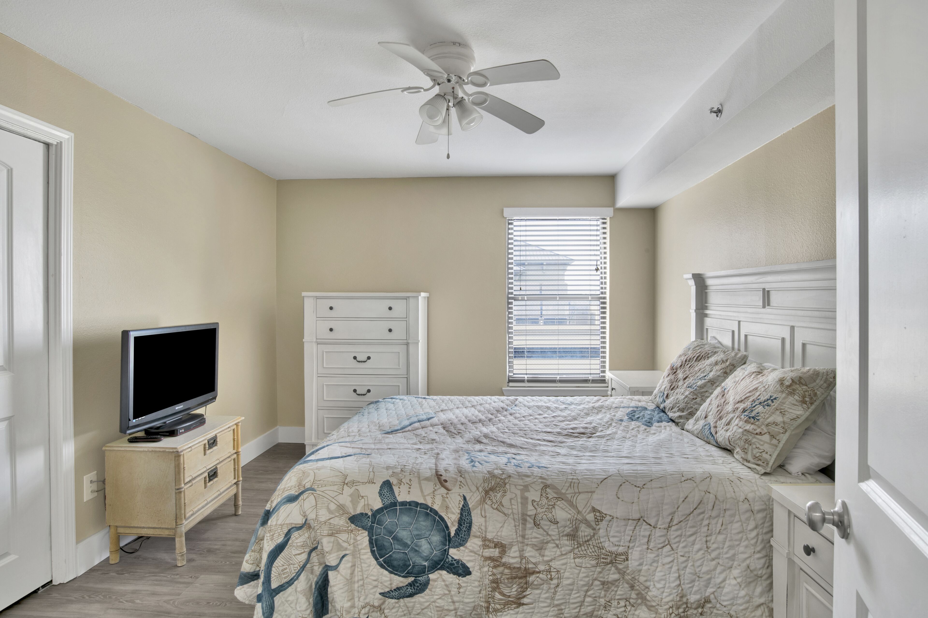 A bedroom with a bed, a ceiling fan, a television on a small table, a dresser, and a window with blinds.