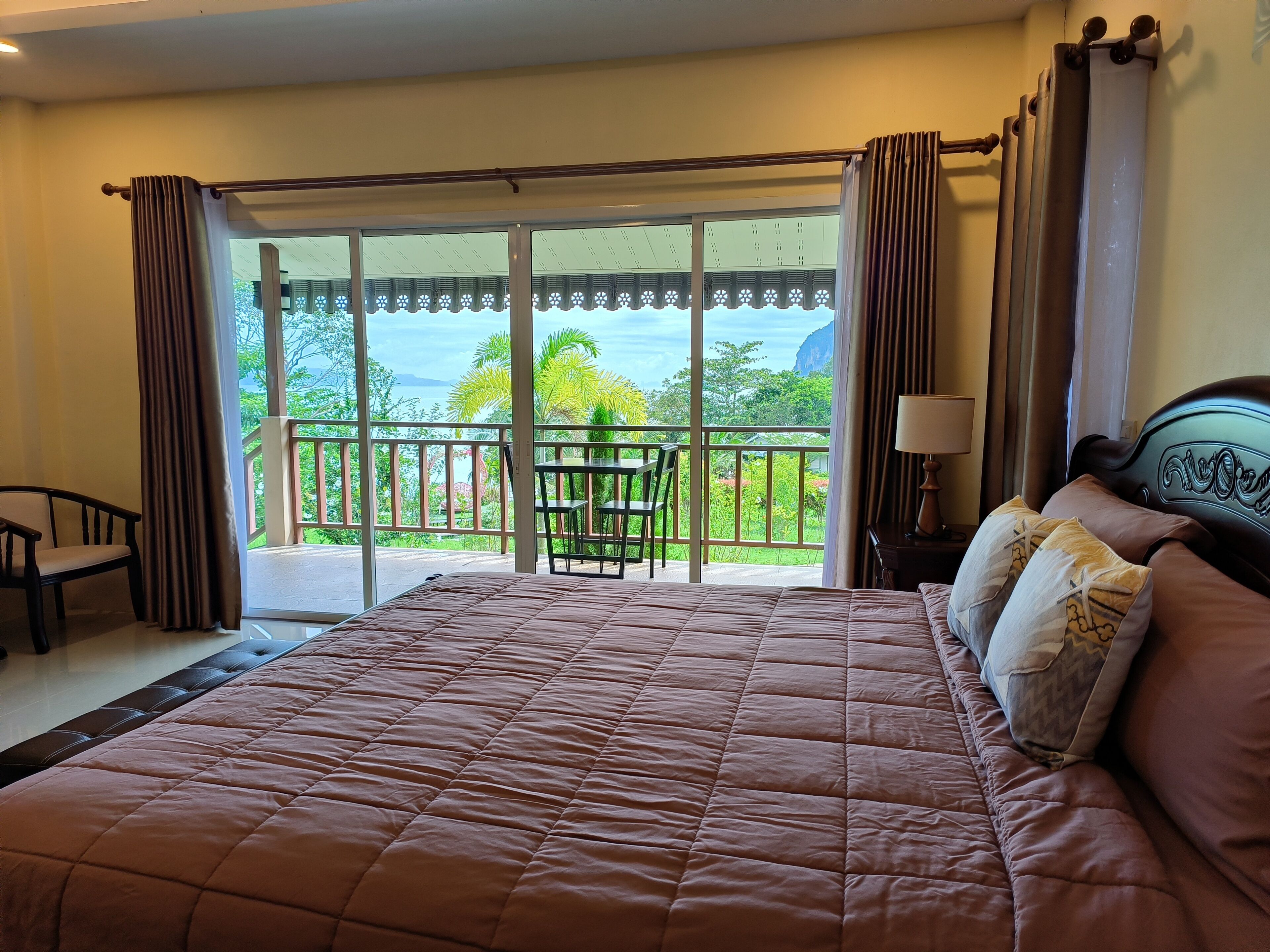 A bedroom with a large bed, a nightstand, a lamp, and a view of a balcony with a table and chairs.