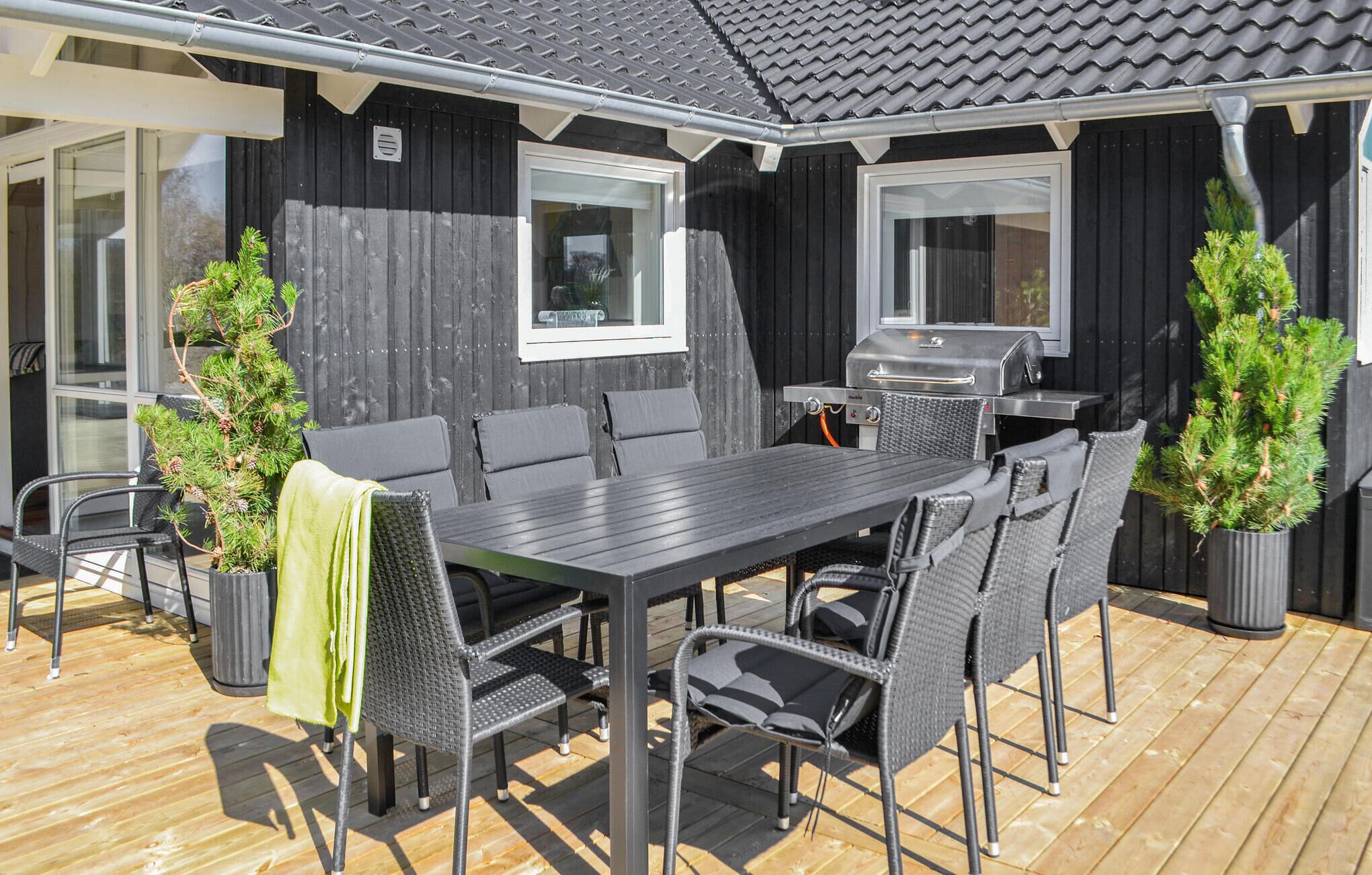 En udendørs patio med en mørk træterrasse, et rektangulært bord med seks stole, og to krukker med vintergrønne planter.