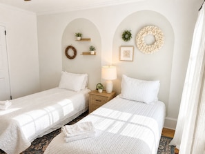 Una habitación con dos camas, una mesita de noche con lámpara, un estante pequeño con plantas y una guirnalda en la pared.