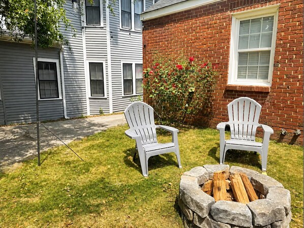 Ein Garten mit zwei Adirondack-Stühlen, einer Feuerstelle aus Stein, einer Ziegelwand und einem Fenster.