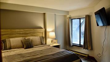 A hotel room with a large bed, a nightstand with a lamp, a sign that says "Last Stop Ski", and a window with curtains.