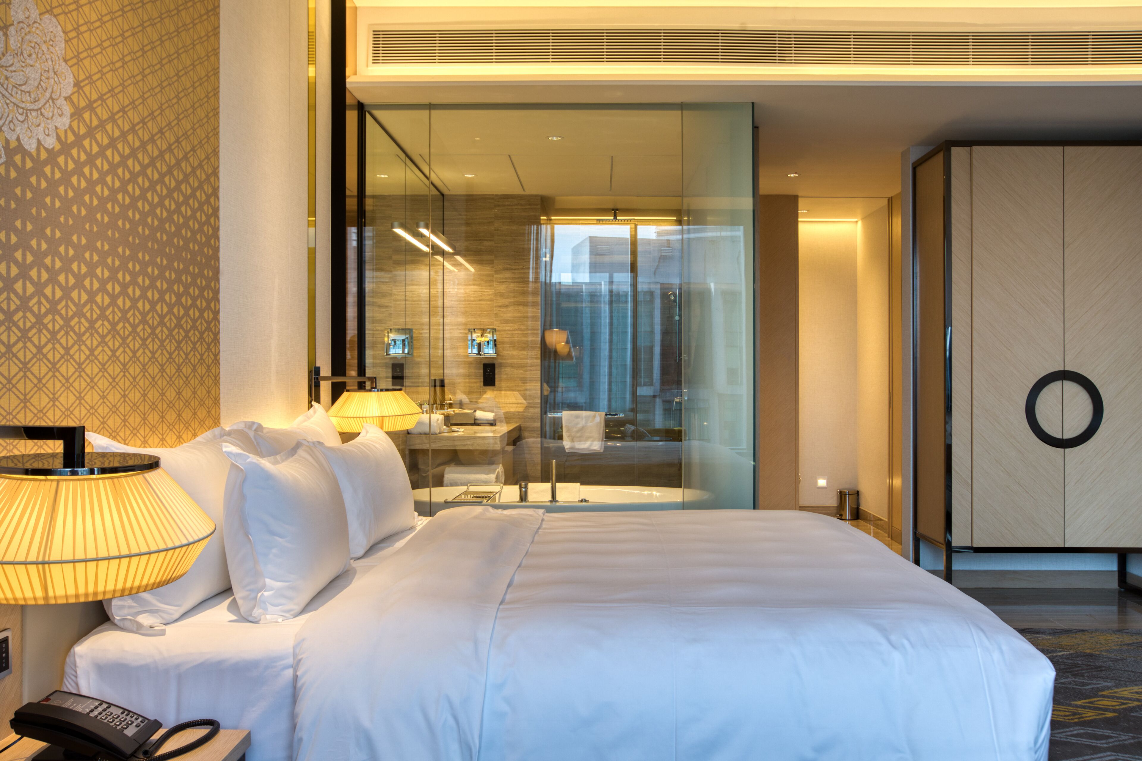 A hotel room with a large bed, a bedside telephone, a glass-enclosed bathroom, and a wardrobe with a circular design.
