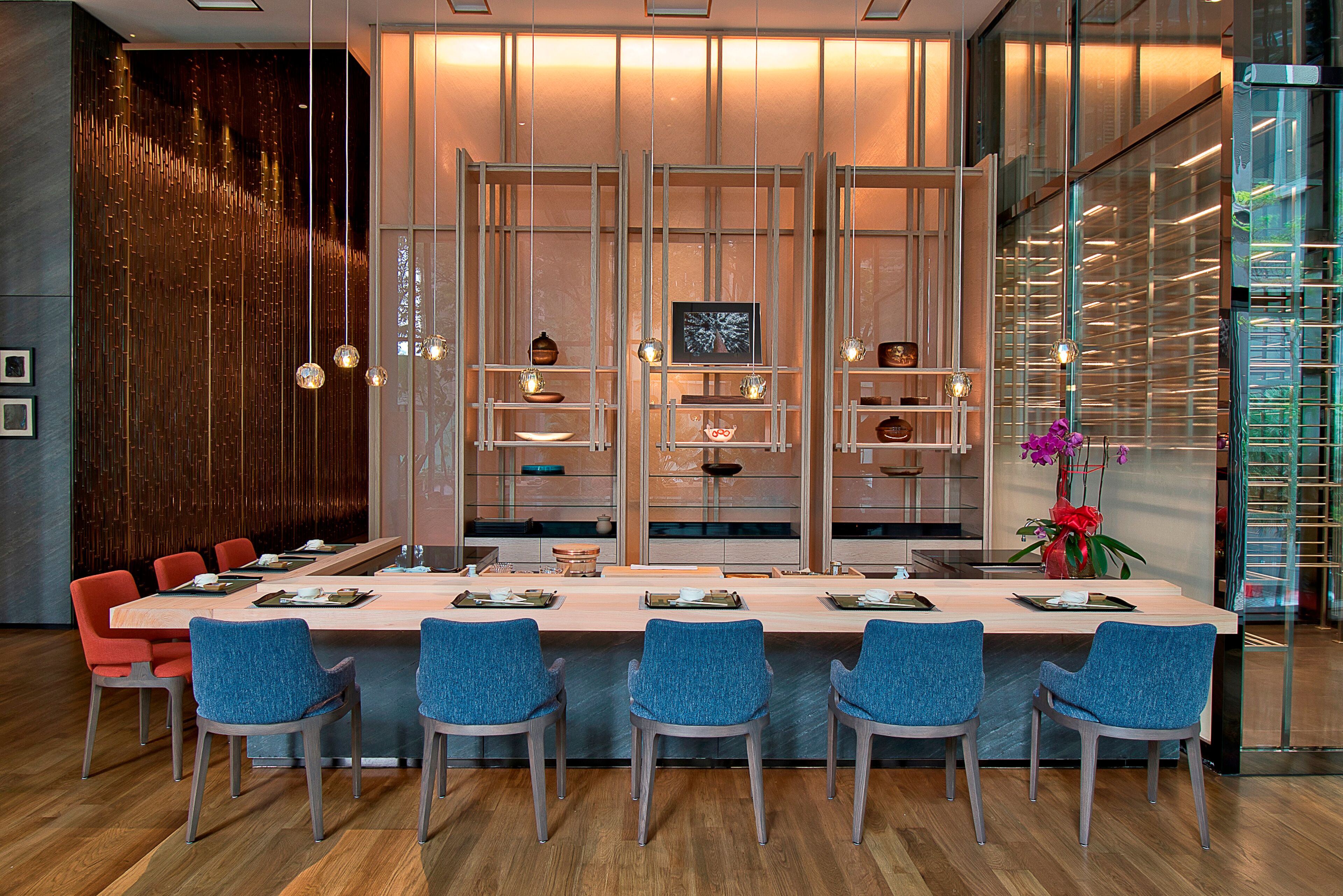 A modern dining area with a long table, blue chairs, and a decorative wall with vertical panels and hanging lights.