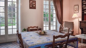 A dining area with a table set for two, a chair, a lamp, and a view through large windows. - Cozy home in Plancoët with WiFi (Plancoët)