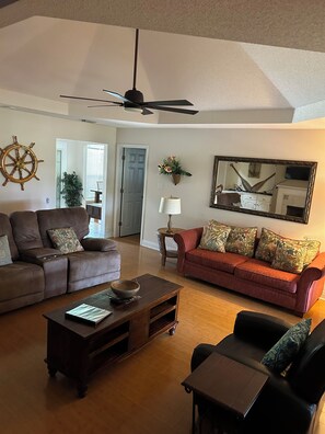 A living room with a brown leather sofa, a red sofa, a wooden coffee table, and a black armchair. There is a ceiling fan, a wall-mounted mirror, and a ship's wheel decoration. - Foley. 20 mins to beaches, close to Tanger Outlet, Owa, sports complex (Foley)