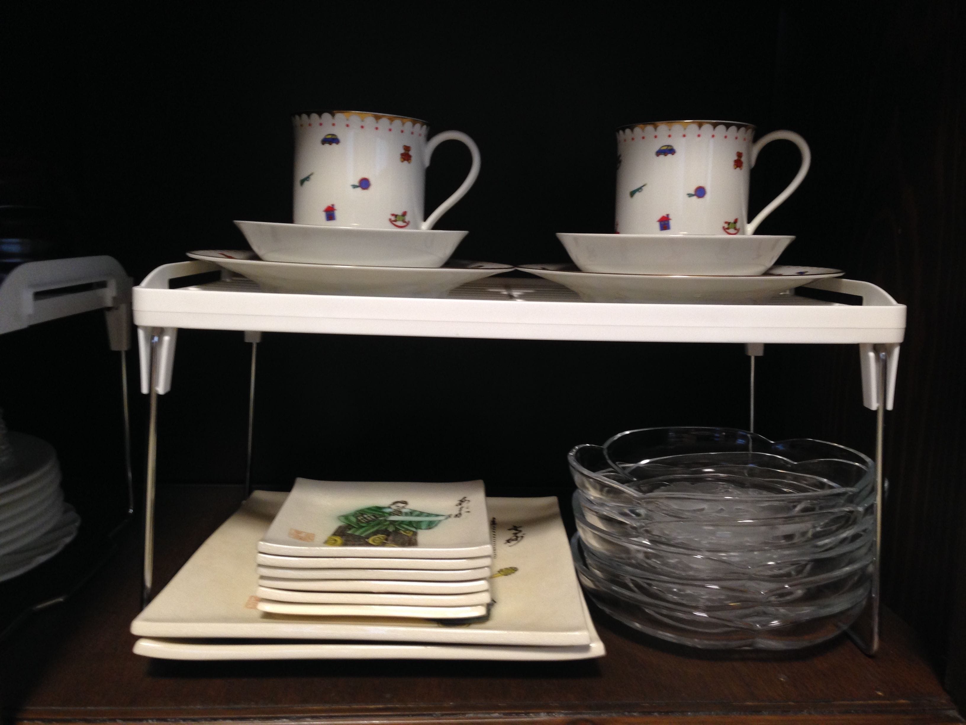 A white tray holding two white cups with colorful patterns and saucers, stacked on a white shelf. Below, there are folded white napkins and a clear plastic container.