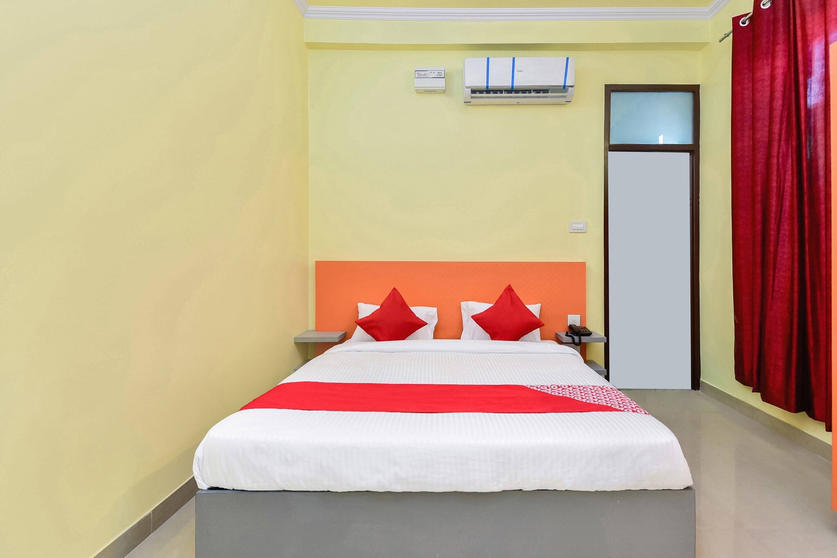 A hotel room with a bed, a nightstand, a red curtain, and an air conditioning unit on the wall.