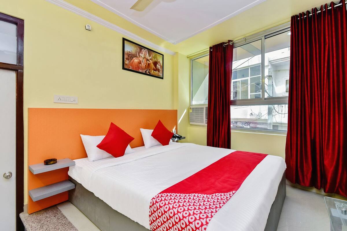 A hotel room with a bed, red curtains, a window, and a painting on the wall.