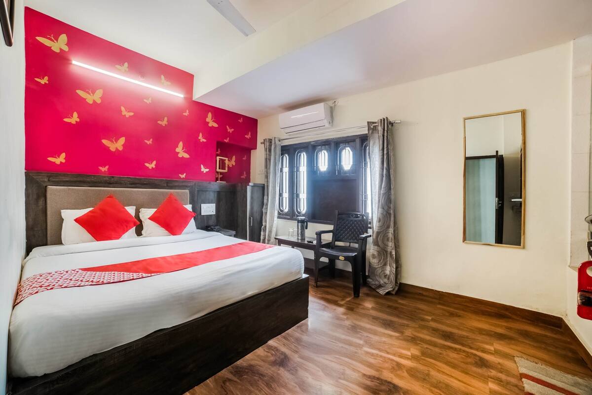 Quarto de hotel com uma cama grande, uma mesa e uma cadeira. Há uma janela com cortinas, um ar condicionado e um espelho na parede.