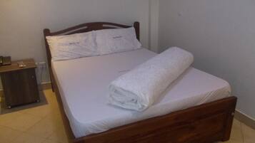 Una cama individual con estructura de madera, sábanas blancas y una toalla enrollada. Se ve una mesita de noche con una lámpara a la izquierda.