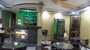 A bar area with a checkered floor, glass tables, and chairs. There is a bar counter with a neon sign, a Heineken refrigerator, and a wall with shelves of bottles. - Rubax Motel (Mbarara)