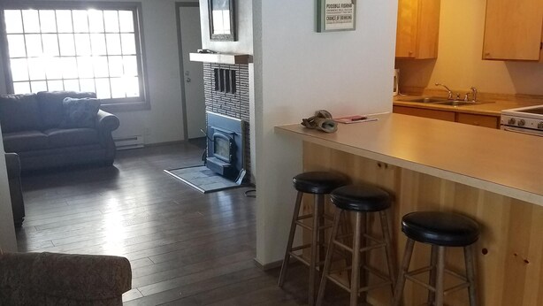 A modern kitchen with a bar area, wooden stools, and a view of the living room. - Little Red Cabin in the woods (Nordman)