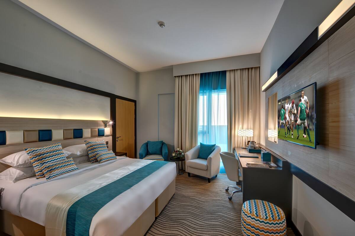 Ein Hotelzimmer mit einem großen Bett, zwei Sesseln, einem Schreibtisch mit Lampe, einem Fernseher und einem Fenster mit Vorhängen.