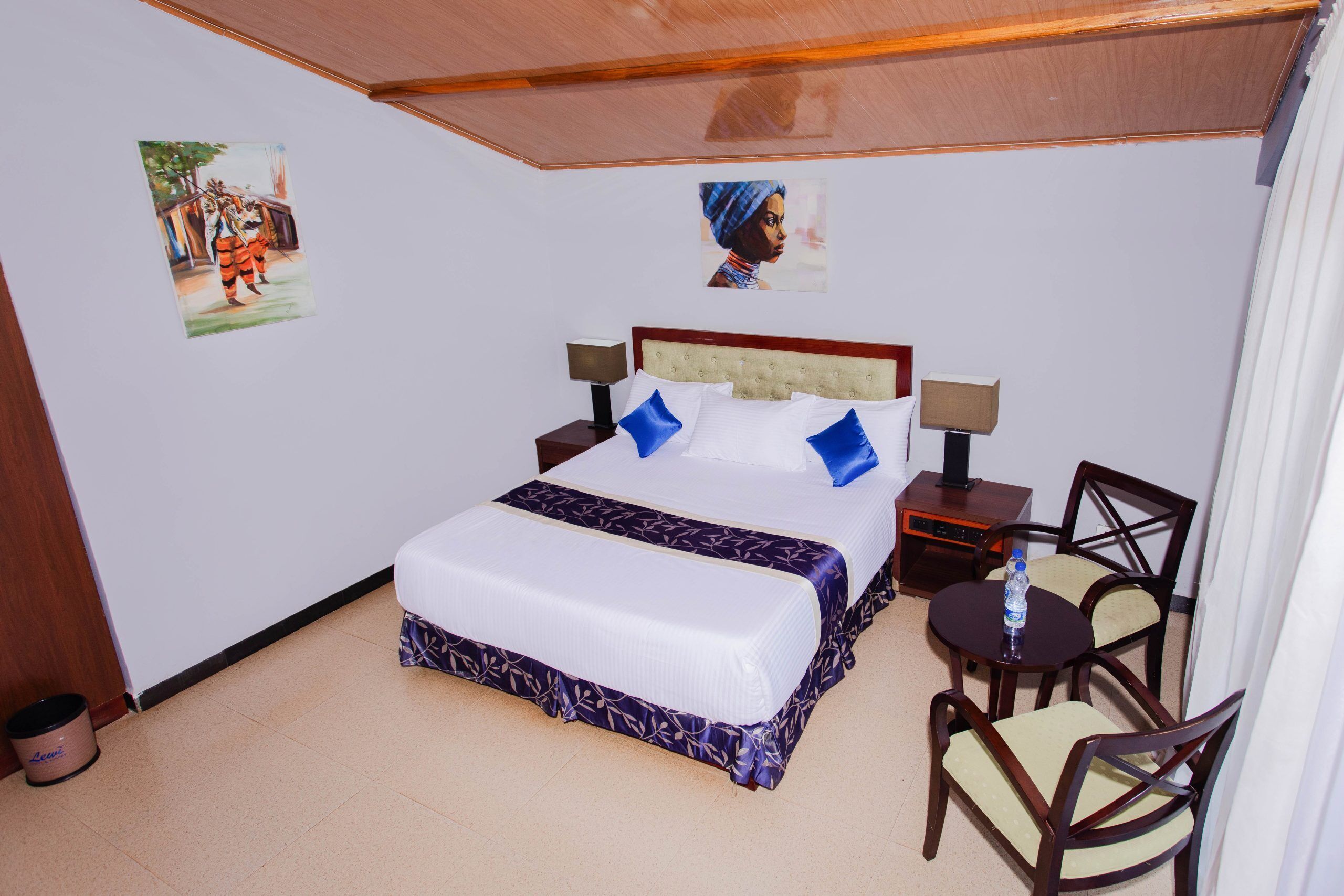 Habitación de hotel con cama, dos mesitas de noche, una silla y una pequeña mesa con una botella de agua.