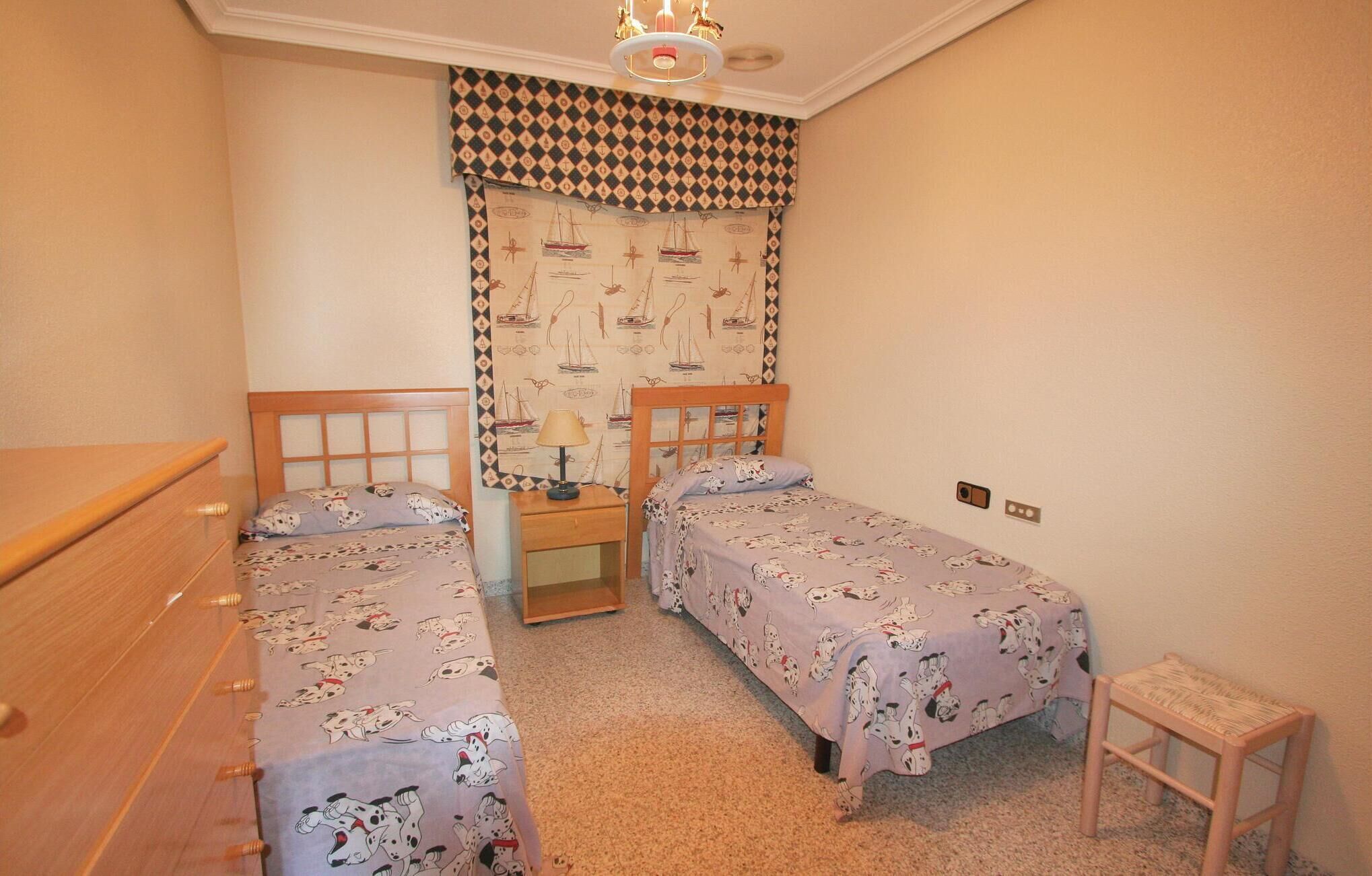Un dormitorio con dos camas individuales, un aparador de madera, una mesita de noche, una lámpara y una cortina estampada.