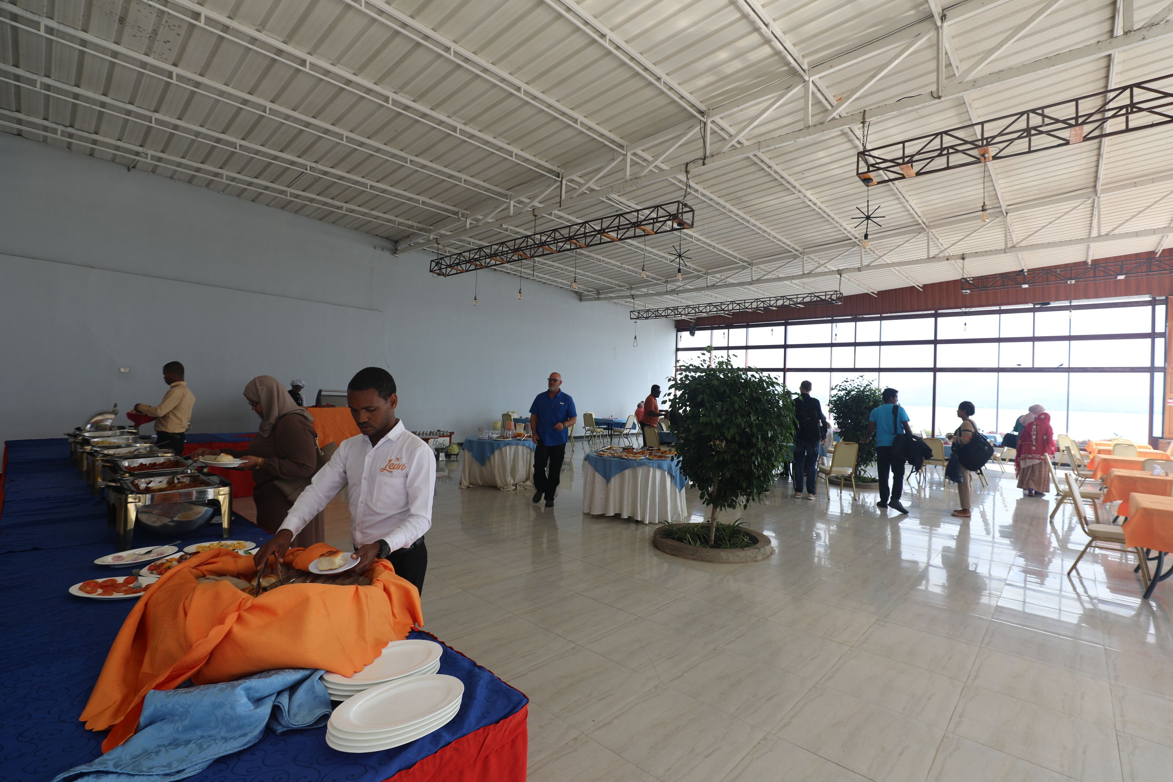 A spacious hall with high ceilings and large windows, featuring a buffet setup with tables and chairs.