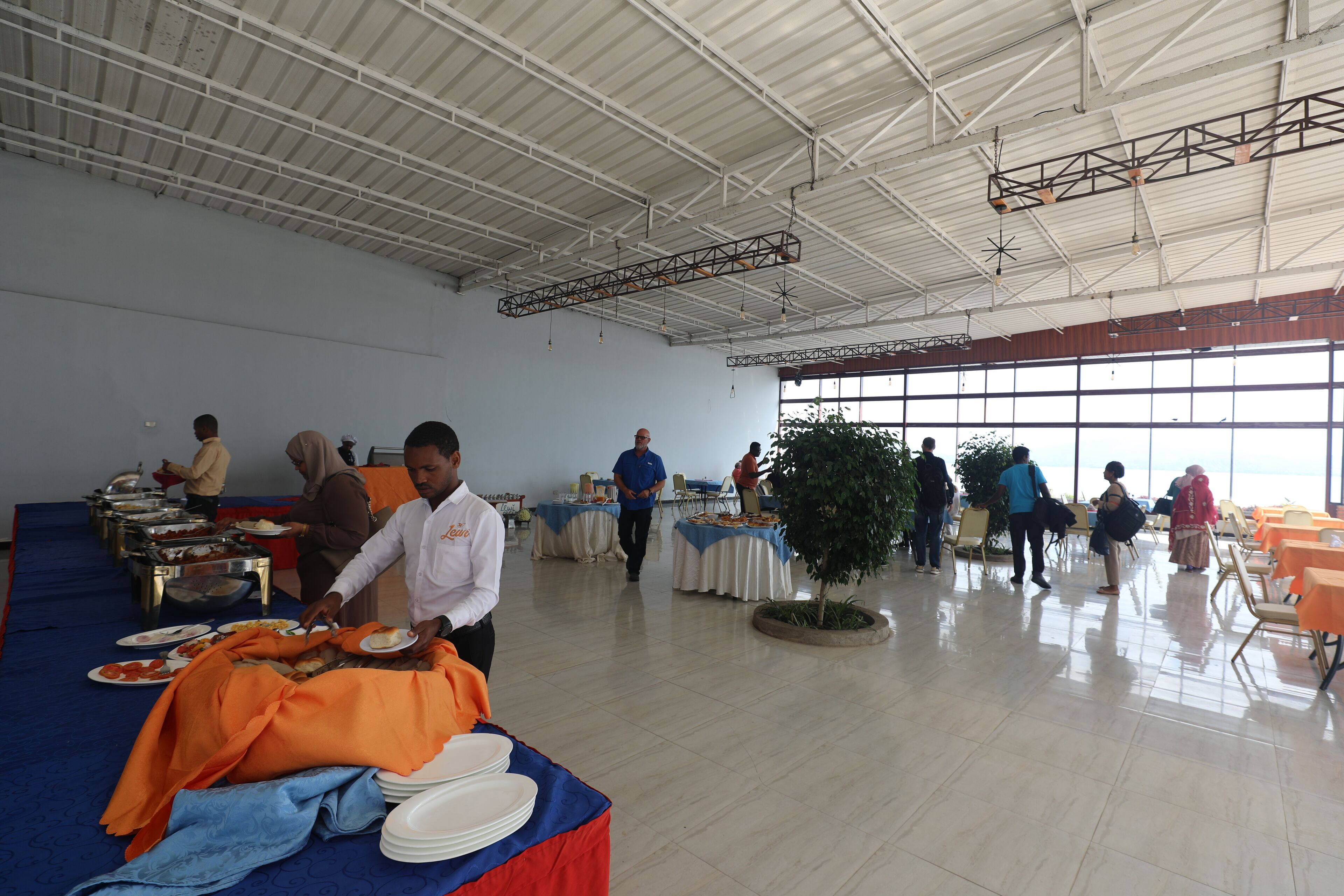A spacious hall with high ceilings and large windows, featuring tables set for a meal with a buffet line visible.