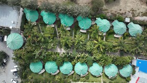 An aerial view of a resort with multiple green-roofed structures, arranged in rows, surrounded by lush greenery and palm trees.