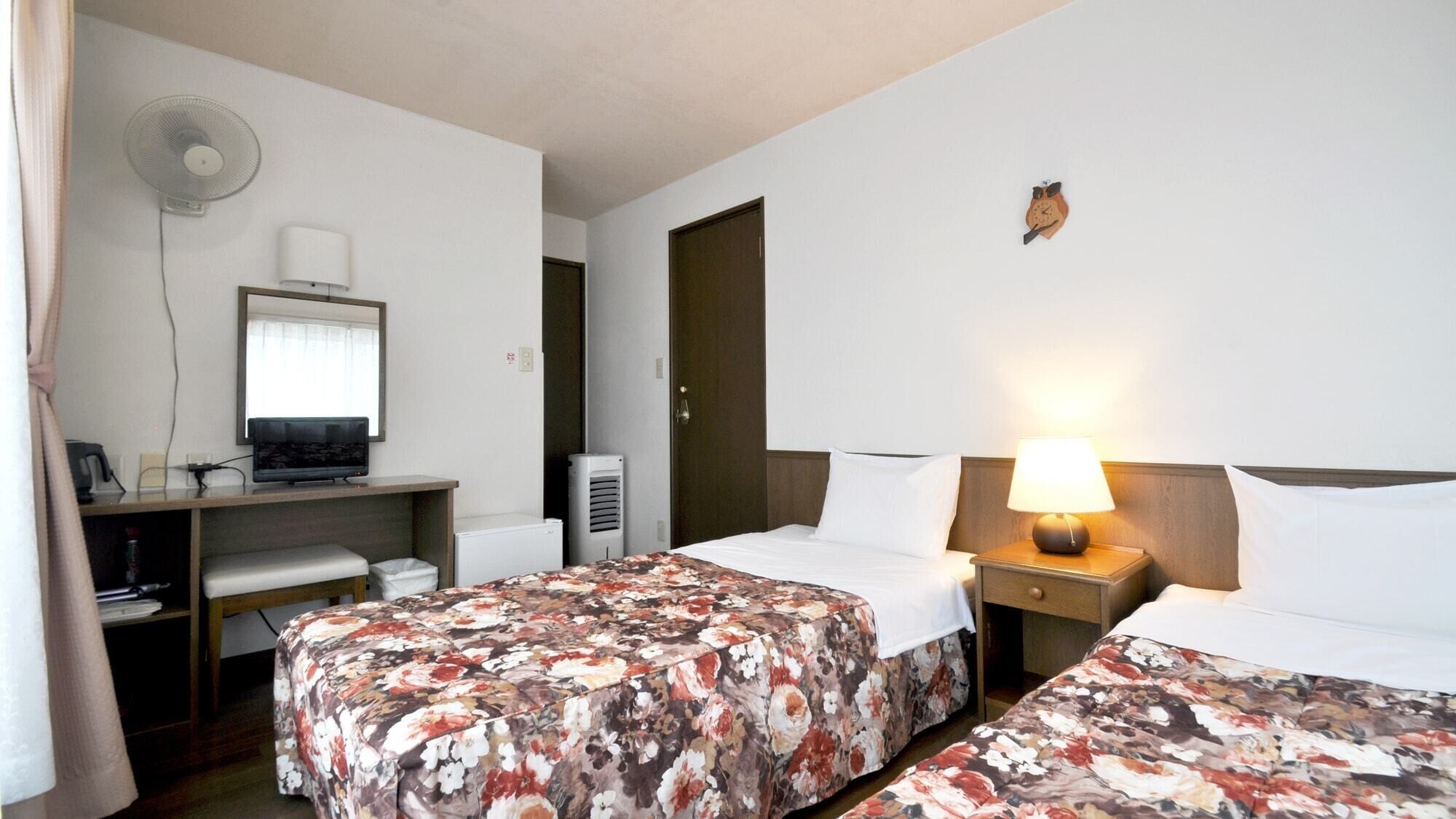 A hotel room with two beds, a desk with a computer, a fan, an air conditioner, and a clock on the wall.