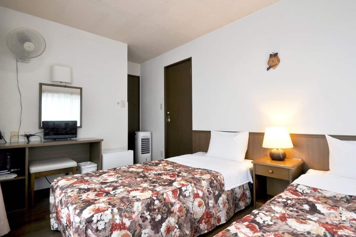 Habitación de hotel con dos camas, un escritorio con computadora, un ventilador, aire acondicionado y un reloj en la pared.