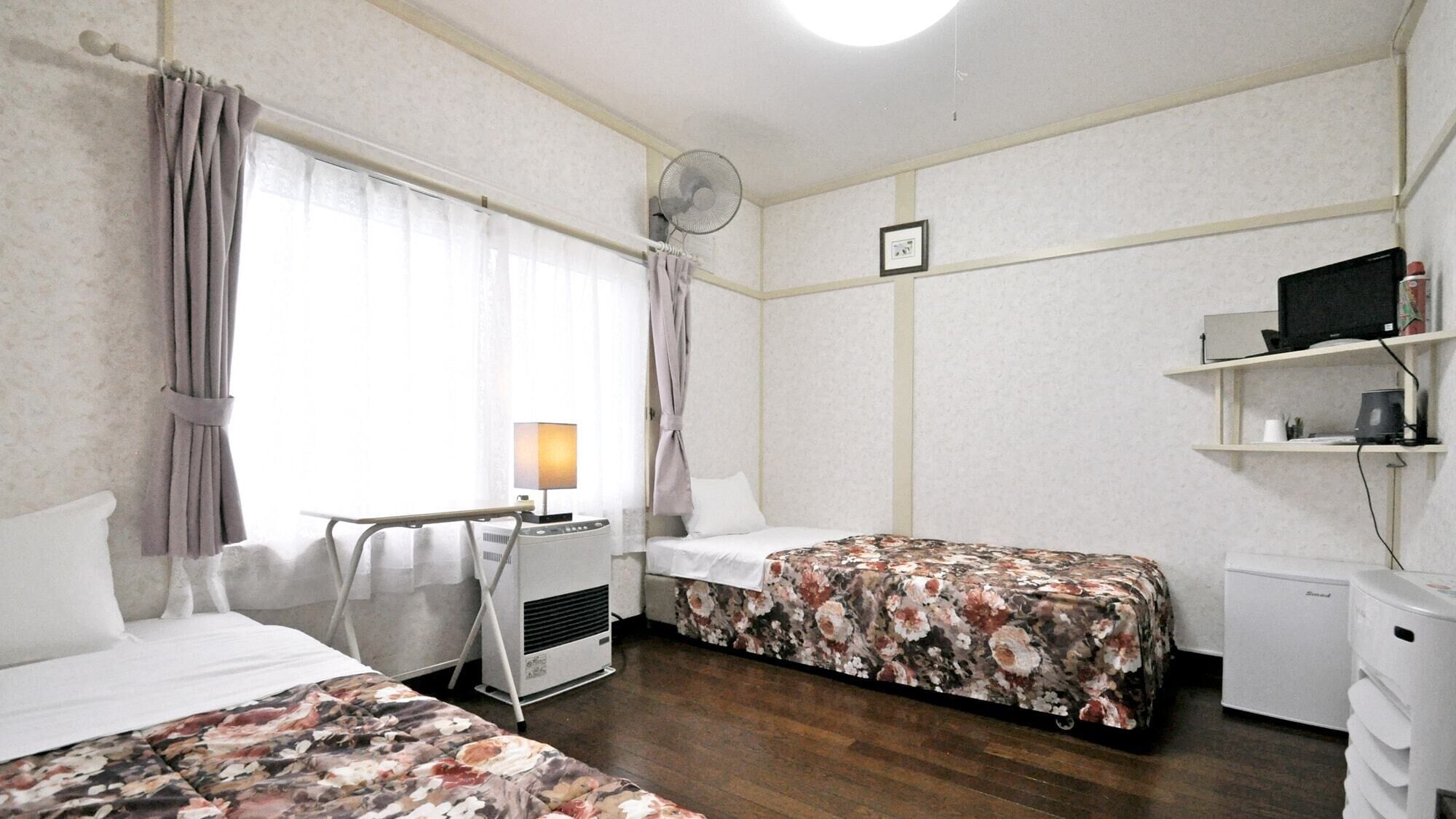 A hotel room with two beds, a desk with a lamp, a small refrigerator, a bedside table, a wall-mounted shelf with a computer, and a ceiling fan.