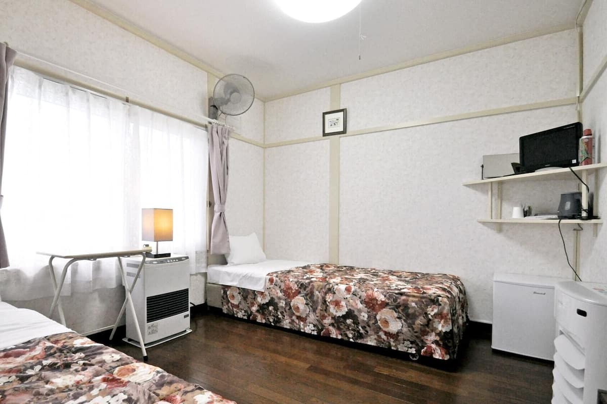 Habitación de hotel con dos camas, un escritorio con lámpara, un refrigerador pequeño, una mesita de noche, un estante en la pared con una computadora y un ventilador de techo.
