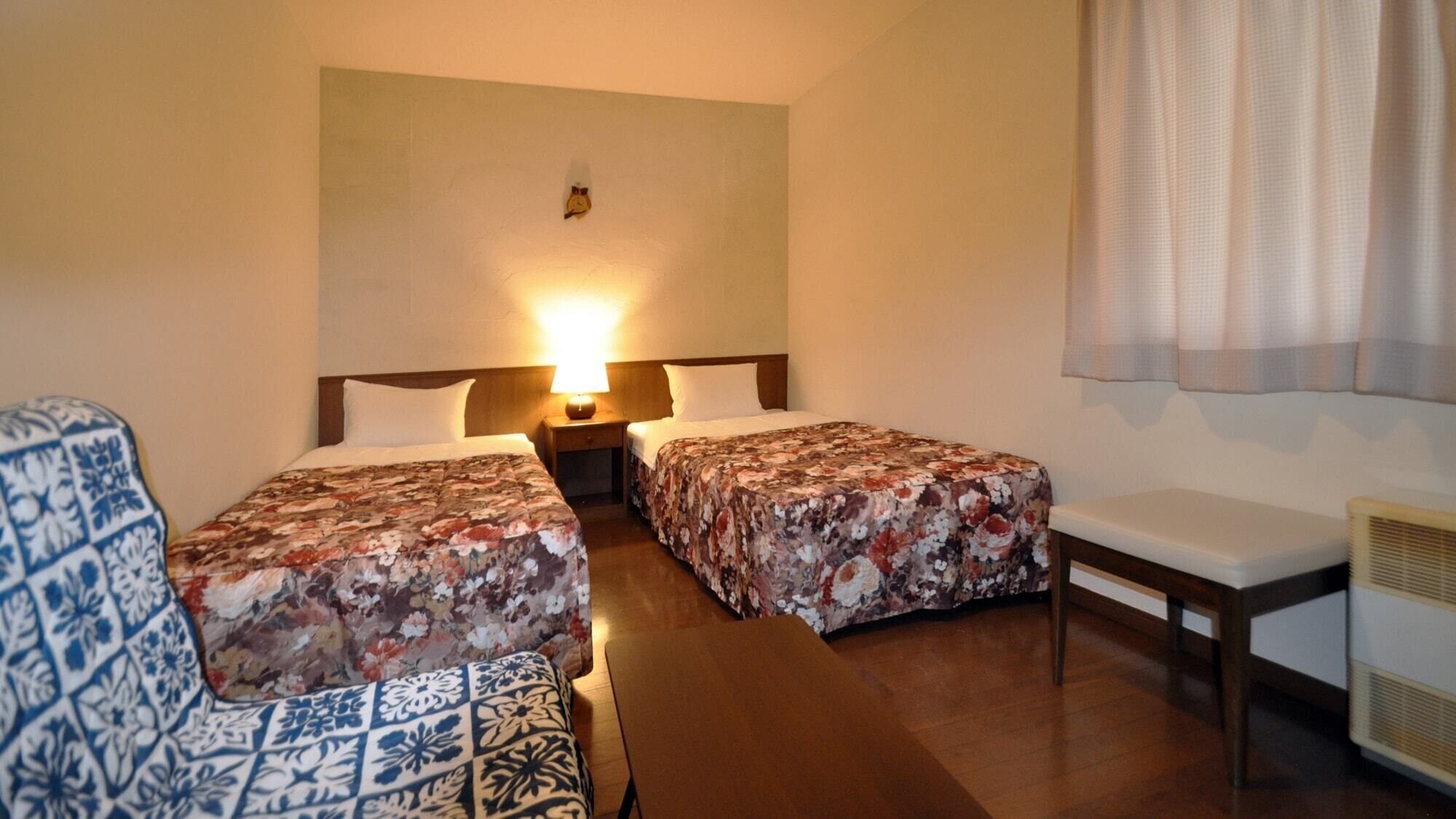 A hotel room with two beds, a small table, a chair, and a heater.