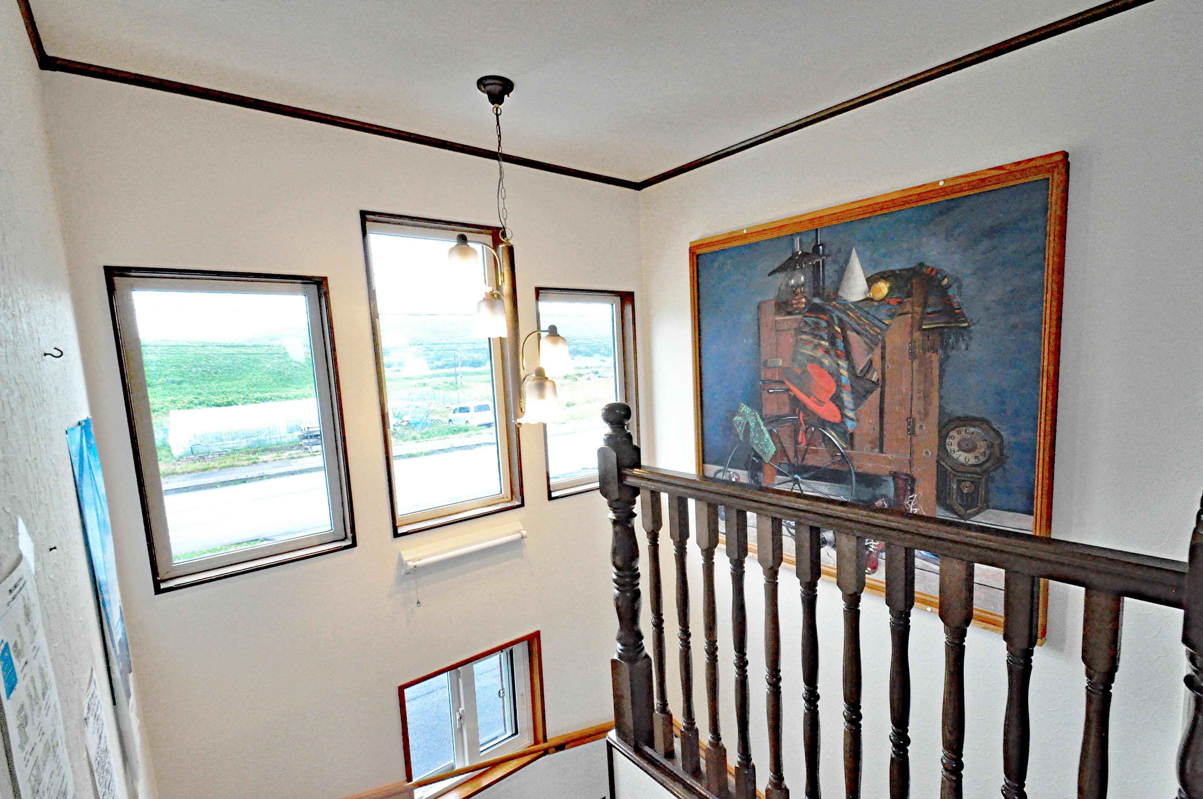 A staircase with a wooden handrail, a painting on the wall, and three windows with views of the outdoors.