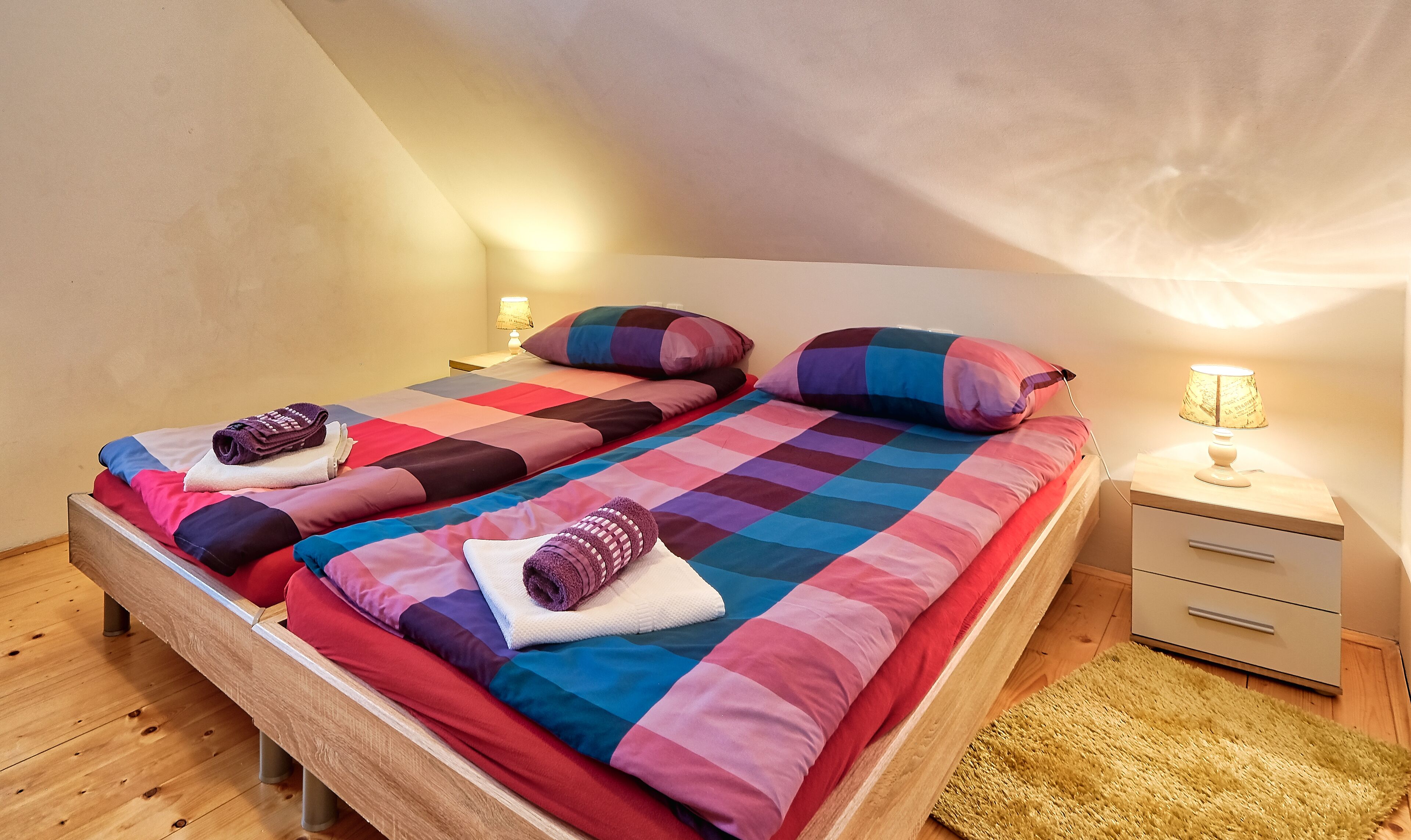 A twin bed with a colorful quilt, two pillows, a bedside lamp, and a wooden nightstand.