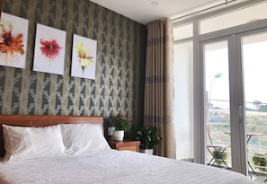 Classic hotel room with a large bed, wooden headboard, and floral artwork, offering a balcony with a view of the surrounding landscape.