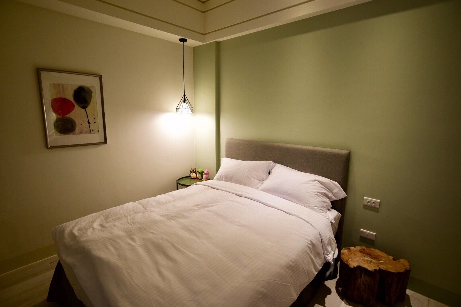 En pænt redt seng i et rum med grønne vægge, et indrammet kunstværk over sengen og et lille natbord med en lampe.