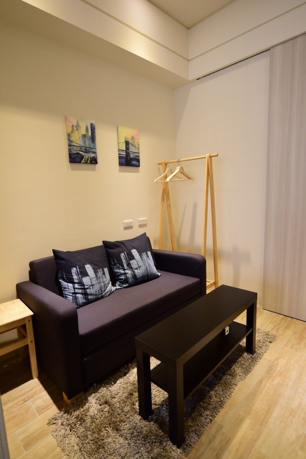 A modern living room with a dark sofa, two black cushions, a wooden hanger stand, a black coffee table, and a light-colored rug.
