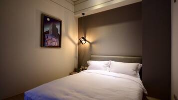 A hotel room with a bed, a nightstand, a wall-mounted lamp, and a painting of a building.