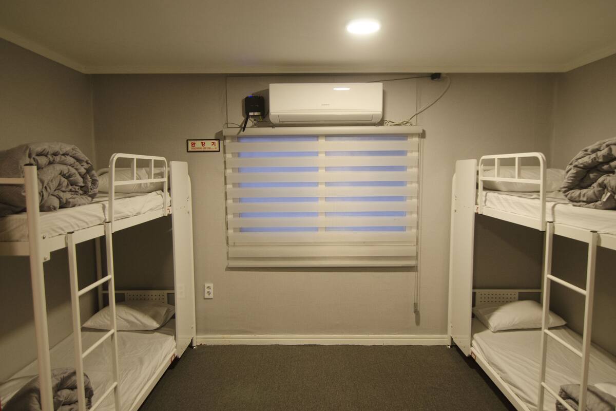 A room with two bunk beds, a window with blinds, an air conditioning unit, and a small framed picture on the wall.