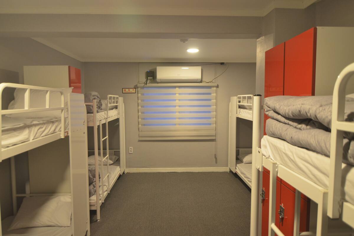 A dormitory room with bunk beds, a window with blinds, an air conditioning unit, and a red cabinet.