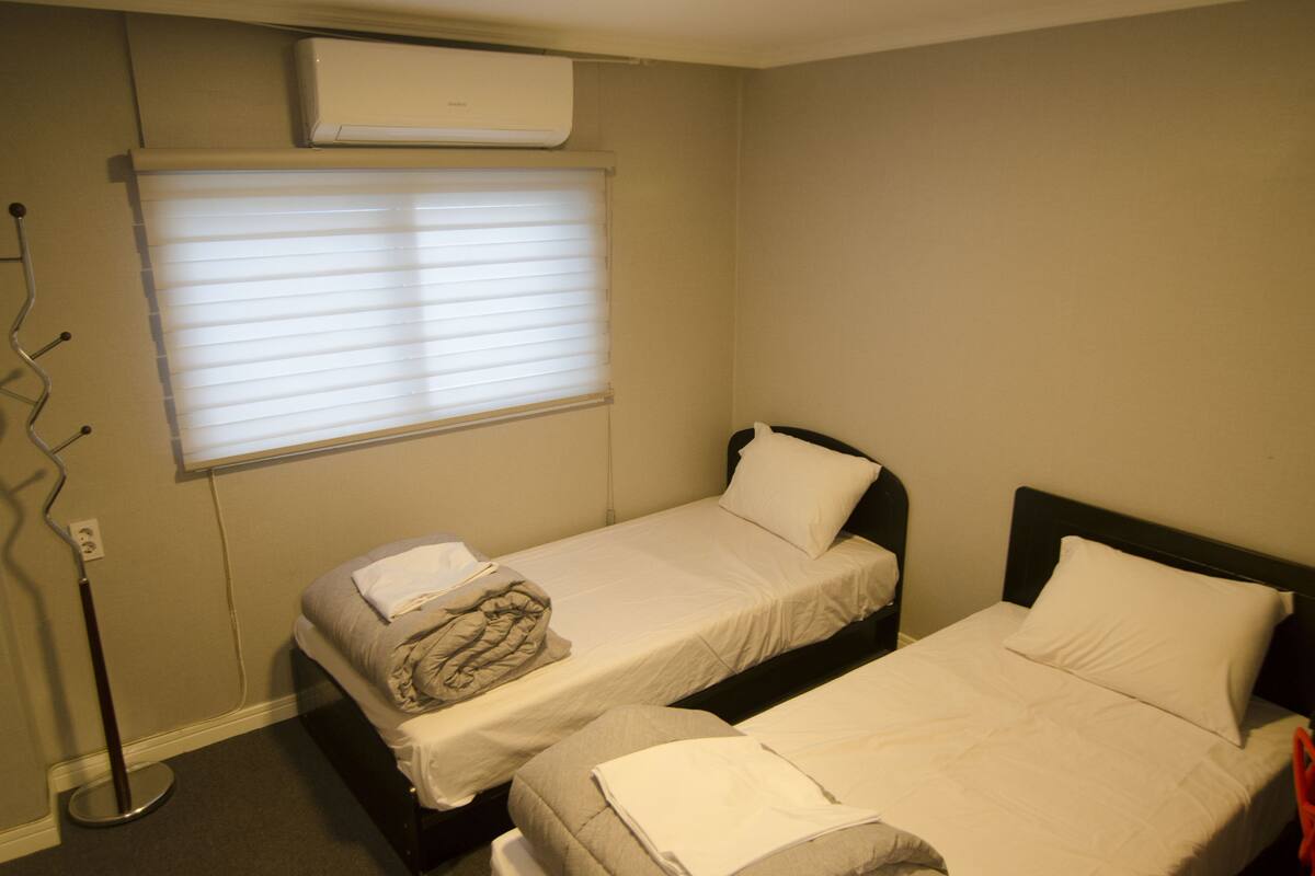 A hotel room with two single beds, a window with blinds, an air conditioning unit, and a floor lamp.