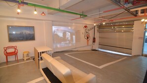 Un espace intérieur doté d’un écran de projection diffusant une vidéo, d’un canapé blanc, d’un fauteuil rouge et d’un petit tabouret.