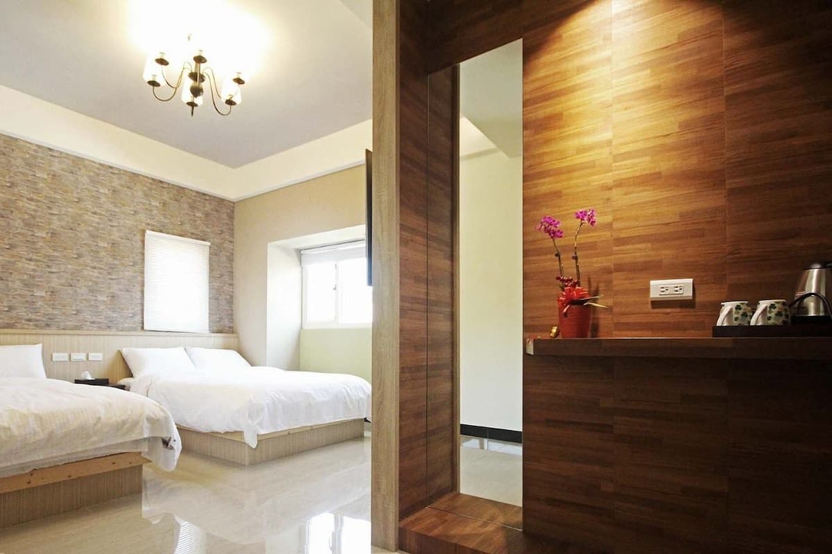 A modern hotel room with two beds, a brick accent wall, a wooden panel wall, and a small table with a vase of flowers.