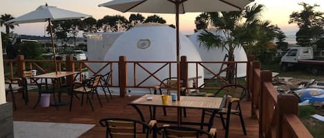 Una terraza al aire libre con mesas y sillas de madera, sombrillas y vistas a una estructura en forma de cúpula.