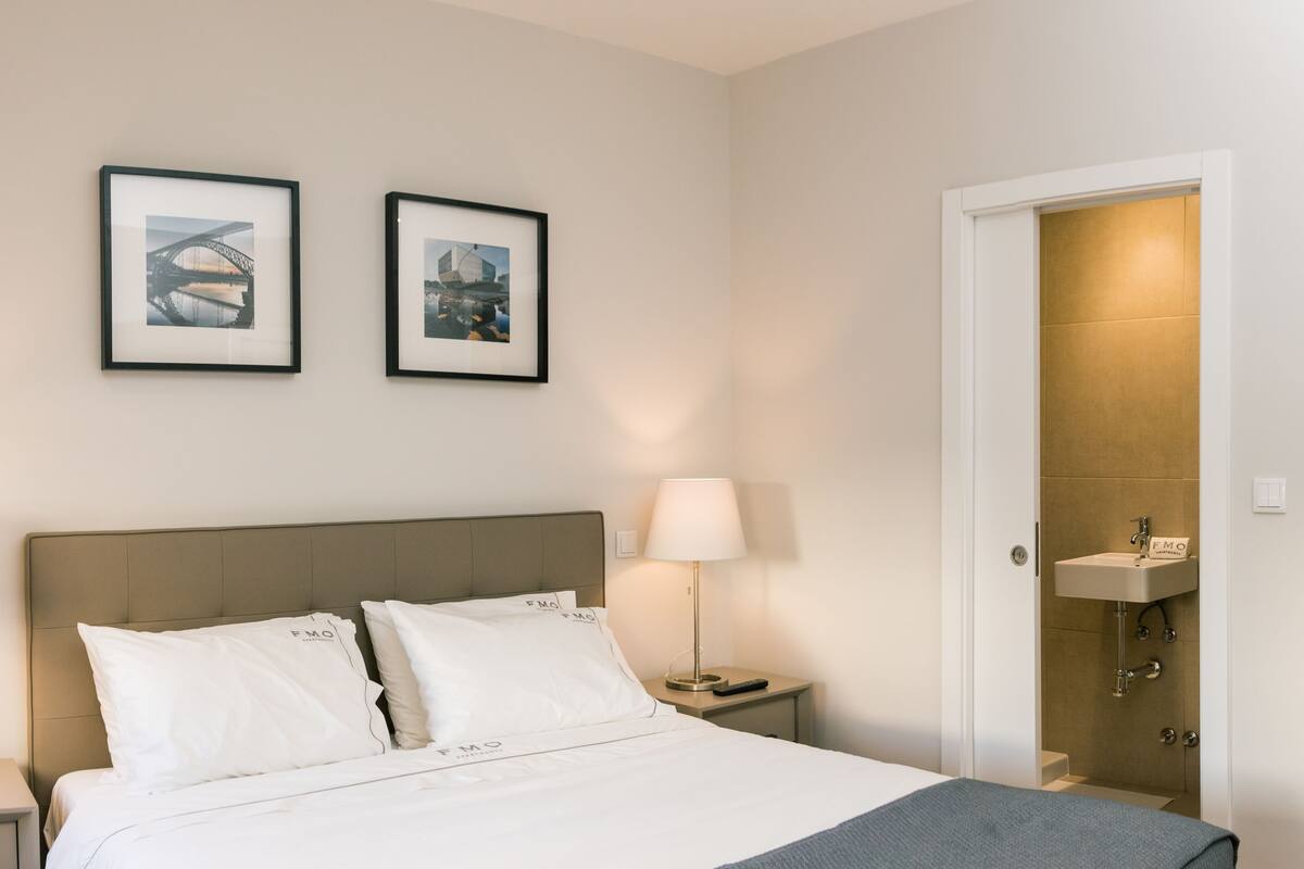 A hotel room with a bed, two bedside tables with lamps, a nightstand, a door leading to a bathroom, and two framed pictures on the wall.