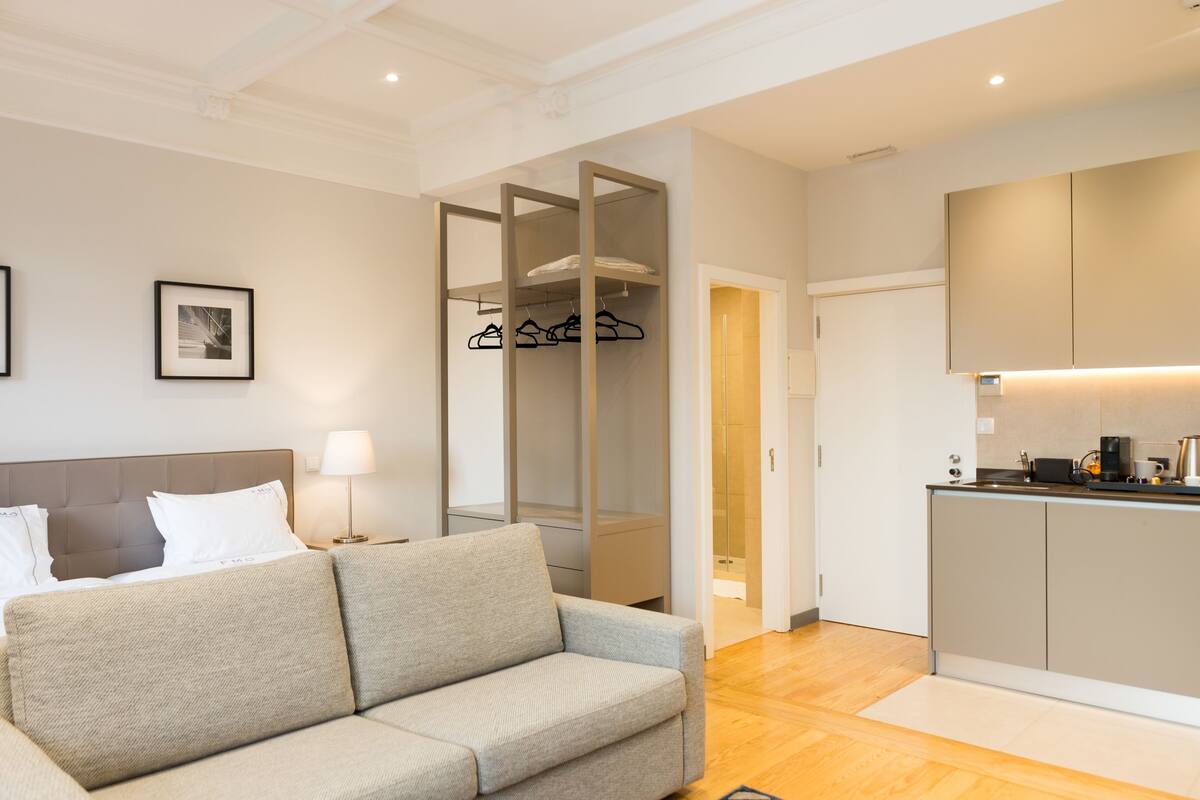 A modern hotel room with a bed, a sofa, a wardrobe, and a kitchenette.