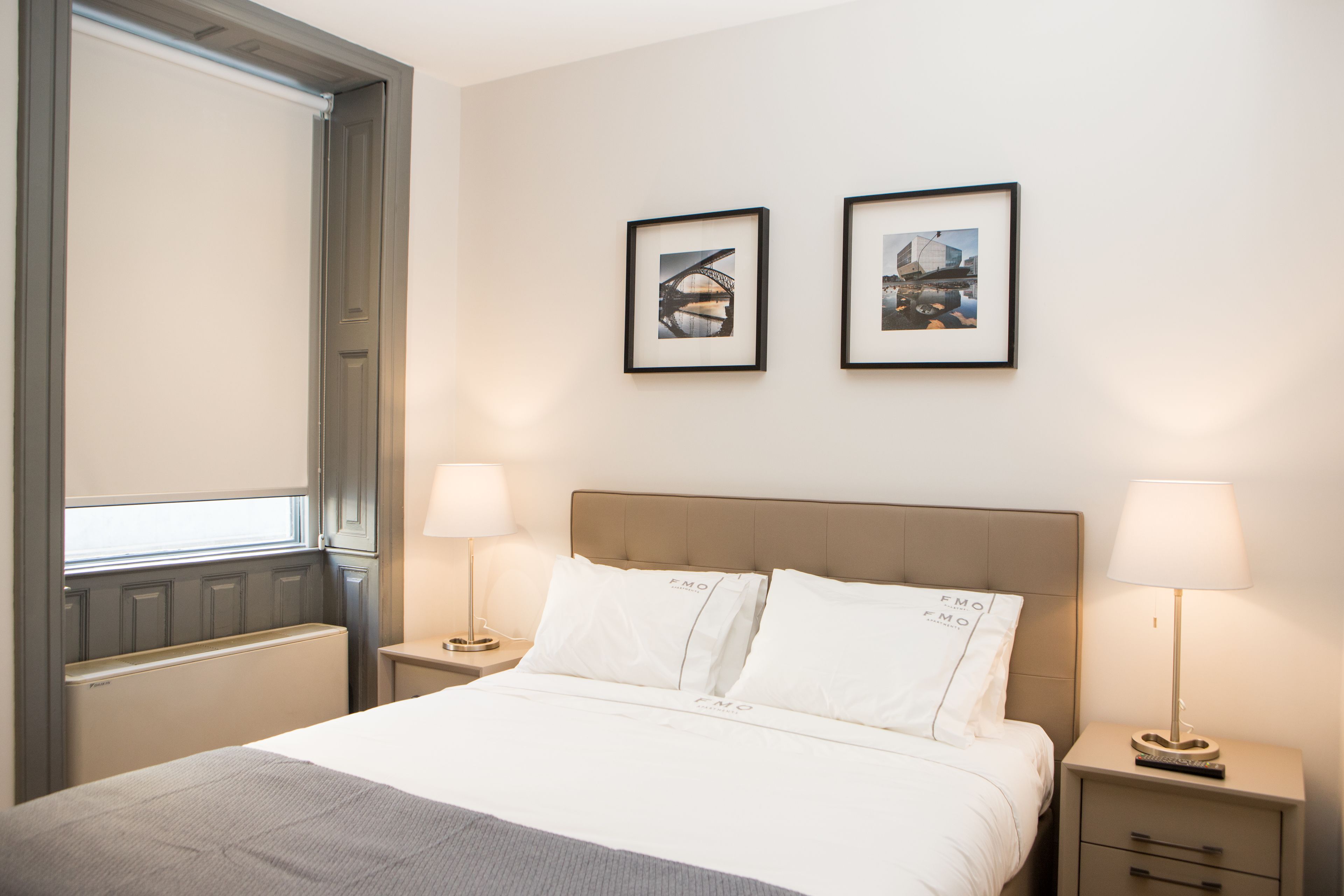 Habitación de hotel con cama, dos mesitas de noche con lámparas, ventana con persianas y dos cuadros en la pared.