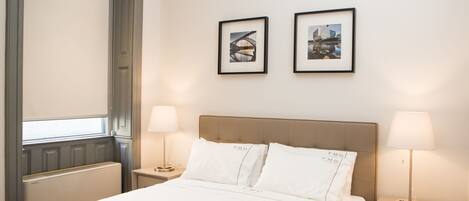 Habitación de hotel con cama, dos mesitas de noche con lámparas, ventana con persianas y dos cuadros en la pared.