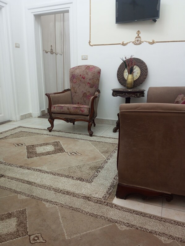 A living room with a floral armchair, a brown sofa, a patterned rug, a decorative clock, and a television on the wall. - New Panorama Hotel (Cairo)