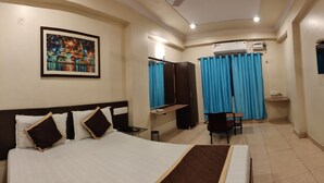 Una habitación de hotel con cama, escritorio, silla, televisión, aire acondicionado y un cuadro en la pared.