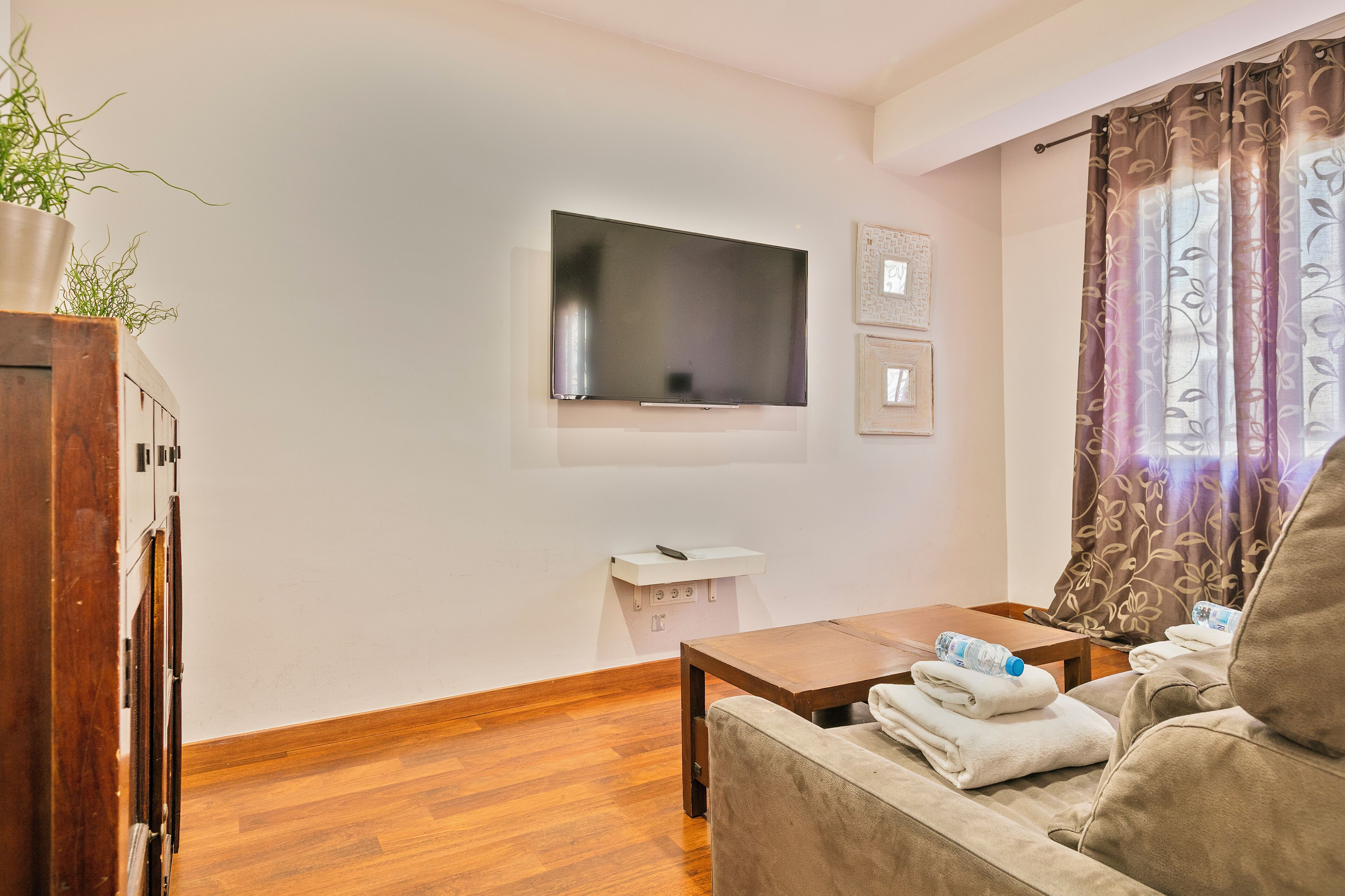 A living room with a flat-screen TV mounted on the wall, a wooden coffee table, a sofa with towels, and a potted plant.
